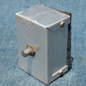 May include: A silver metal box with a black foam pad on one side. The box has a metal knob on the front and two screws on the side.