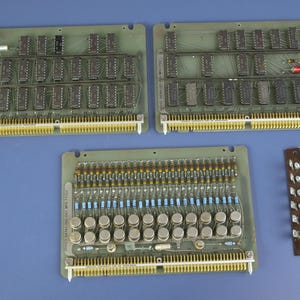 May include: Three rectangular green circuit boards with gold-colored pin connectors along one edge. Two boards feature rows of black integrated circuits, while the third board contains rows of silver metal transistors and small resistors.