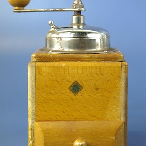 May include: A vintage coffee grinder. It has a light brown wooden body, a metal top and a handle. The wooden body has a small drawer at the bottom. The grinder has a crank handle with a wooden knob.