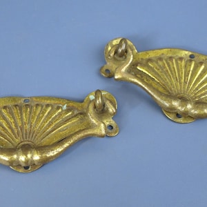 Puede incluir: Dos tiradores de cajón antiguos de latón con diseño en forma de abanico. Los tiradores tienen una forma curvada con un elemento decorativo central y bucles para la fijación. El latón presenta una pátina envejecida.