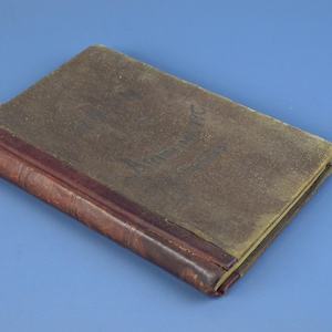 Puede incluir: Un libro antiguo con una cubierta marrón desgastada y un lomo rojo oscuro. El título "Arithmetic" es apenas visible en la cubierta. El libro está cerrado y sobre una superficie azul.