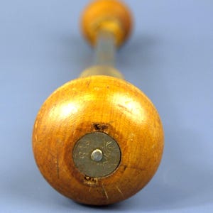 May include: A hand tool featuring a rounded, light brown wooden handle with a circular metal inlay at the base. The metal inlay is engraved with the text 15 and a small symbol. A long, thin metal shaft extends from the center of the handle.