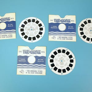 Puede incluir: Carretes y embalajes View-Master vintage sobre un fondo azul. El embalaje es blanco con detalles azules y texto que dice "ONE VIEW-MASTER REEL" y "7 MORE WONDERS OF THE WORLD". Los carretes son blancos con marcos negros.