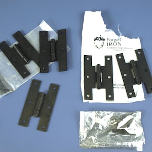 May include: Several black wrought iron H-hinges are displayed on a blue surface. Some hinges are packaged in clear plastic bags with screws. The paper packaging includes the text "ACORN FORGED IRON".