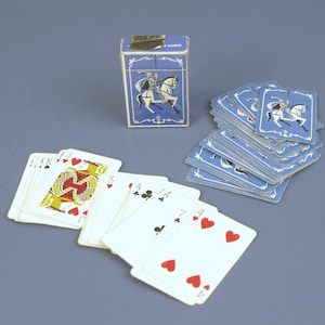 Könnte beinhalten: Ein Vintage-Spielkartenset mit einer blau-weißen Schachtel mit einem Ritter zu Pferd. Das Deck enthält Karten mit roten und schwarzen Farben, darunter Herzen, Kreuz, Karo und Pik. Die Karten sind in einem Fächer und einem Stapel angeordnet.