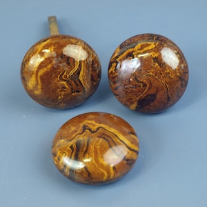 May include: Three decorative cabinet knobs with a marbled pattern in shades of brown and gold. One knob has a brass stem. The knobs are round and have a glossy finish, suggesting they are made of a polished stone or resin.