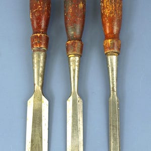 May include: Three vintage Stanley wood chisels with worn, dark red wooden handles and metallic ferrules. Each tool features a steel blade with visible signs of age and use, including surface oxidation and markings on the metal shafts.