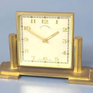 Pode incluir: Um relógio de mesa dourado, em estilo Art Déco, com um mostrador quadrado creme. O relógio tem algarismos pretos e ponteiros dourados. É suportado por dois postes verticais e uma base retangular. O texto "BLACK STAR & FROST" é visível.