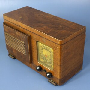 Puede incluir: Una radio vintage de madera con un dial dorado y botones negros. La radio tiene un acabado de madera con veta y una rejilla de altavoz en el lateral.