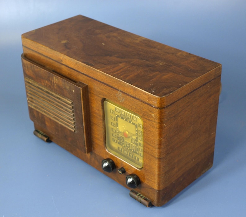 Puede incluir: Una radio vintage de madera con un dial dorado y botones negros. La radio tiene un acabado de madera con veta y una rejilla de altavoz en el lateral.