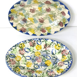 May include: Two oval ceramic platters with hand-painted designs. The top platter has a white base with colourful brushstrokes in yellow, green, and maroon, and a blue scalloped border. The bottom platter has a similar design with blue circles.
