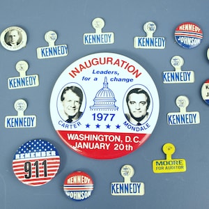 Puede incluir: Una colección de botones y pines de campaña política vintage. El surtido incluye pines con los nombres Kennedy, Johnson, Carter y Mondale. Un botón conmemora los ataques del 11 de septiembre, y otro presenta el texto "Moore for Auditor."