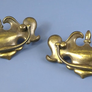 以下が含まれることがあります： 装飾的なデザインの、2つのアンティーク真鍮引き出しプル。各プルは、湾曲したハンドルと、中央のフィニアルとスクロールのようなアクセントを備えた詳細なバックプレートを備えています。プルは、水色の背景に配置されています。