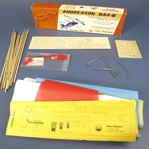 May include: A vintage model airplane kit with a brown and orange box labelled "ANDREASON BA4-B". The kit includes wooden sticks, a red propeller, and yellow instructions. The box reads "Peanut Scale" and "Rubber Powered Flying Scale Models Designed to Fly."