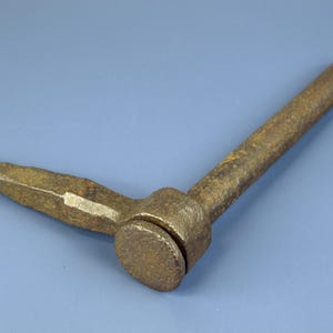 May include: A weathered metal tool, likely antique, with a threaded end and a rounded head. It features a long, cylindrical handle and a shorter, angled arm. The rusty appearance suggests age and use. The tool is probably for mechanical applications.