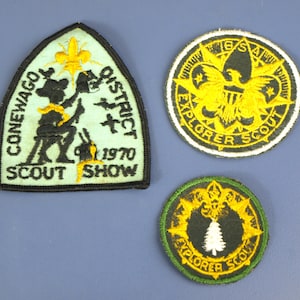 Peut inclure: Trois écussons brodés. Le patch supérieur gauche est vert clair avec le texte noir qui dit "CONEWAGO DISTRICT SCOUT SHOW 1970". Le patch supérieur droit est rond avec du texte jaune et noir qui dit "EXPLORER SCOUT". Le patch inférieur est rond avec un arbre blanc.