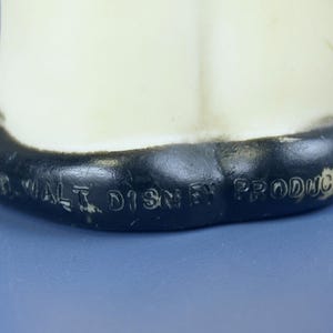 May include: A vintage, off-white figurine with a black base. The base has the text "&copy; WALT DISNEY PRODUCTIONS" embossed in black. The figurine is set against a blue background.