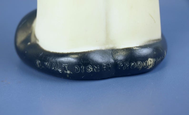 May include: A vintage, off-white figurine with a black base. The base has the text "&copy; WALT DISNEY PRODUCTIONS" embossed in black. The figurine is set against a blue background.