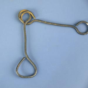 Könnte beinhalten: Ein verwittertes Metallwerkzeug mit einem geschlungenen Griff, einem gebogenen Arm und einem tropfenförmigen Ende. Das Metall hat ein rostiges Aussehen, was auf Alter und Gebrauch hindeutet. Das Werkzeug ist wahrscheinlich für eine bestimmte Aufgabe bestimmt, möglicherweise zum Öffnen oder Manipulieren von etwas.