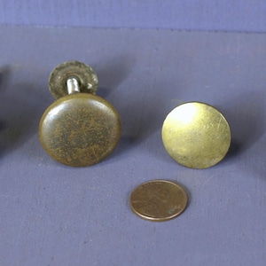 Op de afbeelding: Vier antieke messing kastknopjes met verschillende afwerkingen, waaronder gepolijst, verweerd en aangetast. De knopjes zijn rond en hebben een platte achterkant met een schroefgat. Een penny is meegeleverd ter groottevergelijking.