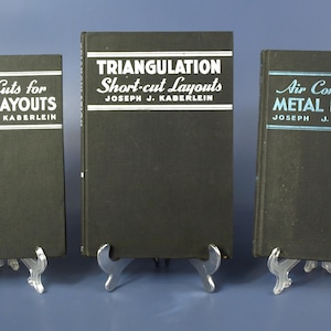 Op de afbeelding: Drie vintage boeken met zwarte covers en witte tekst. De titels zijn "Short Cuts for Round Layouts", "Triangulation Short-cut Layouts" en "Air Conditioning Metal Layout". Elk boek is van Joseph J. Kaberlein.