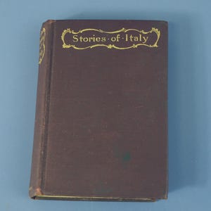 May include: A dark brown hardcover book titled Stories of Italy with gold lettering on the front cover and spine. The title is enclosed in a decorative gold border. The book has a rectangular shape and a solid, matte-textured cover surface.