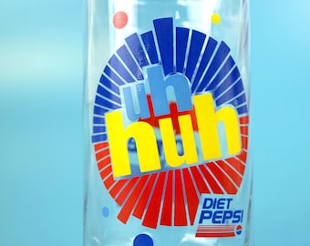 Può includere: Bicchiere trasparente Diet Pepsi con un logo colorato. Il logo presenta le parole "uh huh" in giallo e blu, circondate da elementi grafici rossi e blu. Il bicchiere è su uno sfondo azzurro.