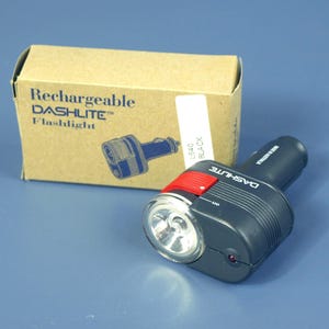 Puede incluir: Una linterna recargable DASH LITE negra con un botón de encendido rojo y una lente transparente. La linterna está junto a una caja de cartón con el texto "Rechargeable DASH LITE Flashlight".