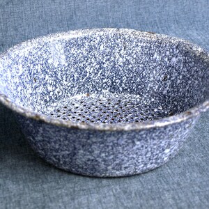 May include: A speckled blue and white enamelware colander. The vintage kitchenware features a round shape with a perforated base and a rolled rim. The colander is designed for draining liquids from food items.