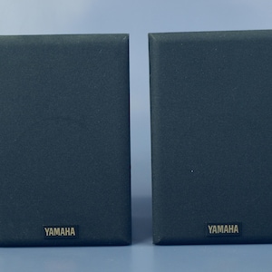 Puede incluir: Dos altavoces Yamaha de color gris oscuro. Cada altavoz tiene forma rectangular con una textura similar a la tela. El logotipo de Yamaha es visible en la parte inferior de cada altavoz. Los altavoces están diseñados para sistemas de audio.