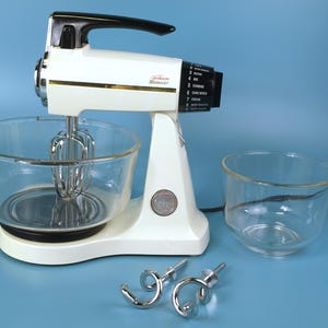 Peut inclure: Un batteur sur socle vintage Sunbeam Mixmaster blanc avec une poignée noire et un cadran de contrôle de vitesse. Il comprend un grand bol mélangeur en verre fixé à la base, un petit bol en verre séparé et deux crochets pétrisseurs en métal chromé.