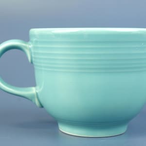 Puede incluir: Una taza de té de cerámica turquesa con base redondeada y asa curva. La taza presenta detalles horizontales estriados alrededor de la parte superior. La taza está sobre un fondo azul liso.
