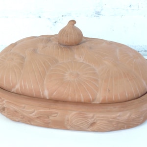 May include: An oval terracotta baking dish with a lid, decorated with carved onion shapes. The lid has an onion-shaped knob. The base of the dish features a carved leaf design. The dish is approximately 25 cm long.
