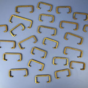 Puede incluir: Colección de pequeñas piezas de ferretería metálicas en forma de C, de color latón, dispersas sobre una superficie azul claro. La ferretería tiene una barra rectangular con extremos curvos, probablemente para muebles.