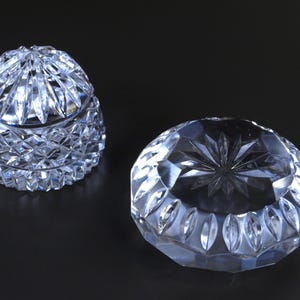 May include: Two clear crystal decorative pieces. One is a lidded, rounded jar with a diamond-cut pattern. The other is a round, open dish with a starburst design in the center. Both pieces are on a dark background.