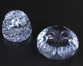May include: Two clear crystal decorative pieces. One is a lidded, rounded jar with a diamond-cut pattern. The other is a round, open dish with a starburst design in the center. Both pieces are on a dark background.