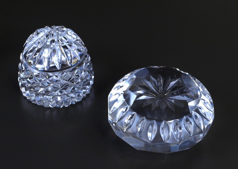 May include: Two clear crystal decorative pieces. One is a lidded, rounded jar with a diamond-cut pattern. The other is a round, open dish with a starburst design in the center. Both pieces are on a dark background.
