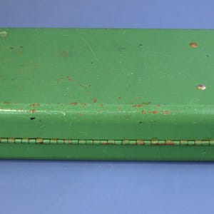 May include: A rectangular, vintage green metal box with a hinged lid. The box has a slightly distressed appearance with visible wear and tear, including rust spots. The box is closed and sits on a blue surface.