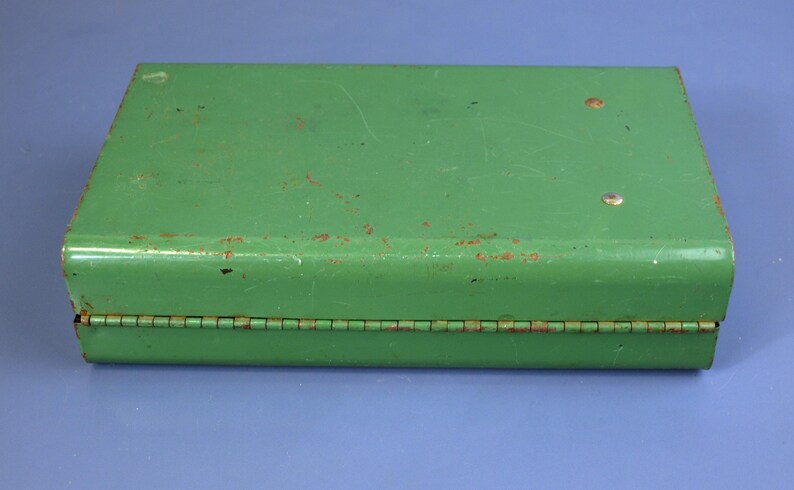 May include: A rectangular, vintage green metal box with a hinged lid. The box has a slightly distressed appearance with visible wear and tear, including rust spots. The box is closed and sits on a blue surface.