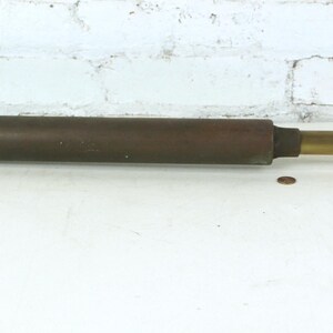 May include: A vintage metal hand pump featuring a long, cylindrical dark brown body with a T-shaped handle at one end and a brass-colored sliding piston rod extending to a flat, wide metal foot plate at the opposite end for manual operation.