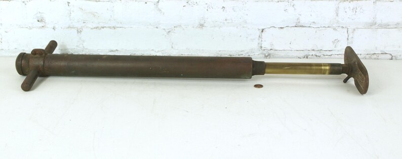 May include: A vintage metal hand pump featuring a long, cylindrical dark brown body with a T-shaped handle at one end and a brass-colored sliding piston rod extending to a flat, wide metal foot plate at the opposite end for manual operation.