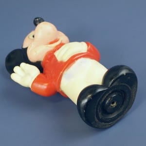 May include: A vintage Mickey Mouse figurine, lying on its back. The figure has a black head and ears, a pink face, and a red jacket. The hands are white, and the shoes are black. The figurine is made of plastic.