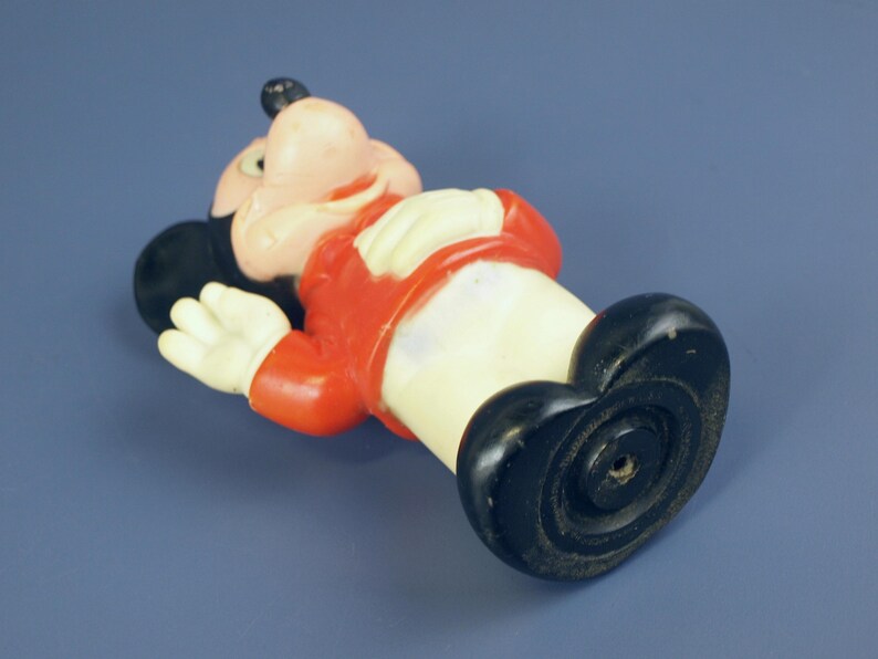 May include: A vintage Mickey Mouse figurine, lying on its back. The figure has a black head and ears, a pink face, and a red jacket. The hands are white, and the shoes are black. The figurine is made of plastic.