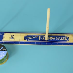 Könnte beinhalten: Ein hölzernes Deluxe E-Z Bow Maker Werkzeug mit einer langen rechteckigen Basis mit blauen und beigen Markierungen, zwei vertikalen Holzdübeln und einem Spulenhalter-Etikett. Daneben steht eine runde Spule mit grünem Offray-Band.