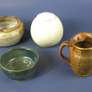 May include: Four ceramic pieces: a small open bowl, a lidded sphere, a small bowl, and a pitcher with a handle. The pieces are in shades of brown, green, and cream, with speckled and glazed finishes. The items are handmade.