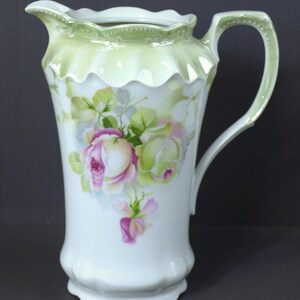 Puede incluir: Una jarra de porcelana blanca con un diseño floral verde y blanco. La jarra tiene un borde festoneado y un asa.