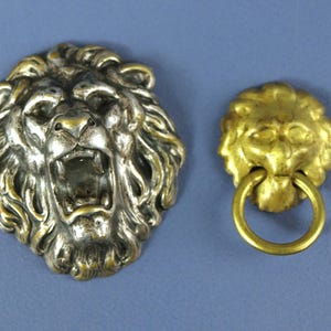 May include: Two metal lion head decorative pieces. One is silver-toned with an open mouth, the other is gold-toned with a ring handle. Both are detailed with manes and facial features, set against a blue background.