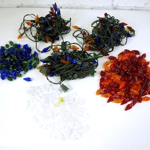 Puede incluir: Una colección de luces navideñas en varios colores, incluyendo azul, verde, naranja y rojo. Algunas luces están ensartadas en cables verdes, mientras que otras están sueltas. Las luces están dispuestas sobre una superficie blanca.