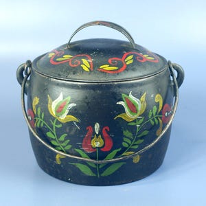 Puede incluir: Un recipiente de metal negro con tapa y asa, adornado con diseños florales pintados a mano en rojo, amarillo y verde. El recipiente tiene un asa metálica y dos bucles laterales para un asa más grande. El diseño es rústico y vintage.