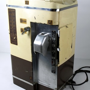 May include: Vintage cream and brown vending machine with a silver metal front panel. The machine has a hinged top and a coin slot. The text "To Operate" is visible on the side. The machine has a power cord.